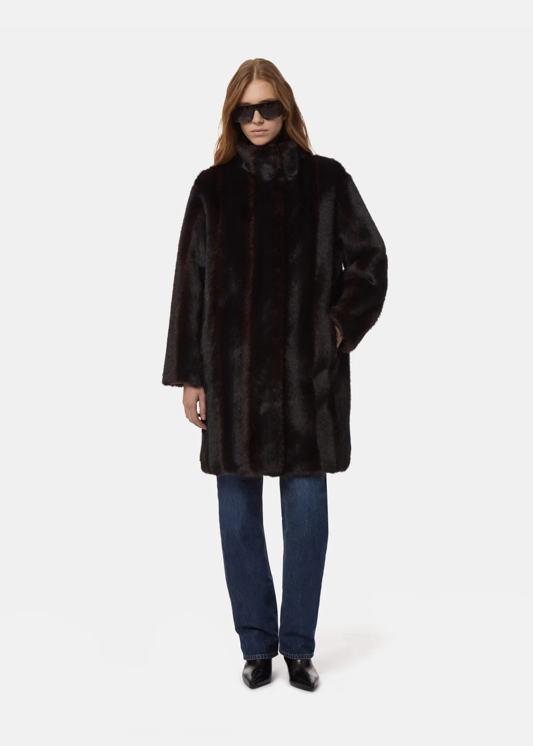 A model wearing a dark brown fur coat, blue jeans, and black sunglasses, standing against a plain light gray background.