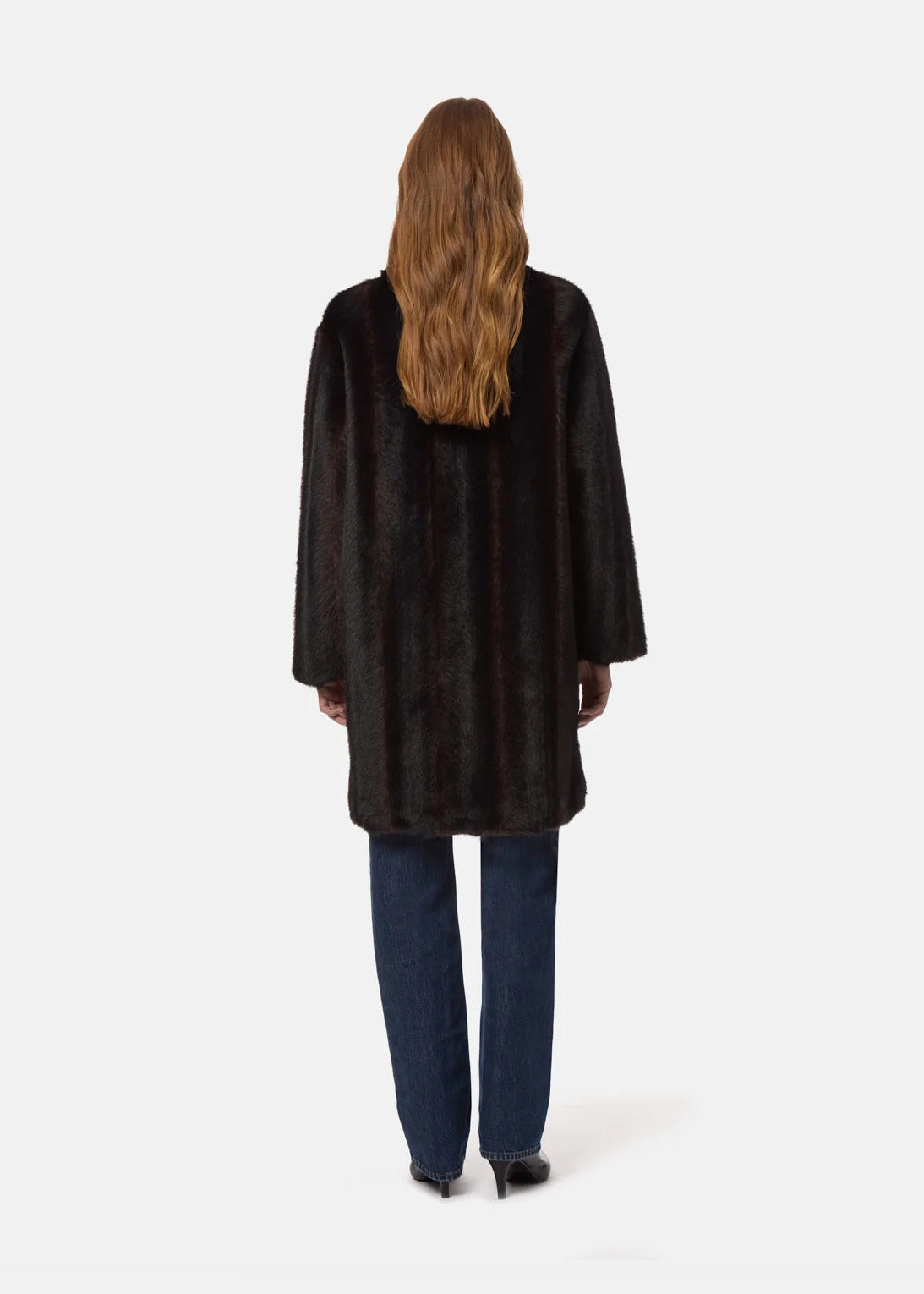 A woman viewed from behind wearing a black faux fur coat, blue jeans, and black heels against a plain white studio background.