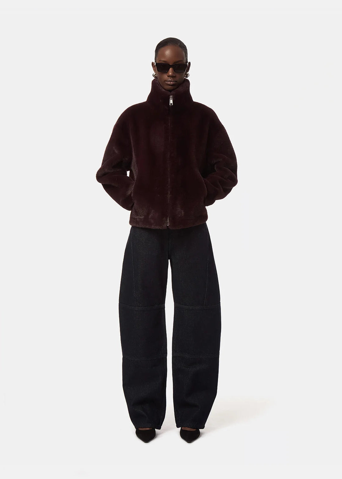 A model wearing a dark burgundy plush zip-up jacket and black wide-leg trousers, standing against a light gray studio background.