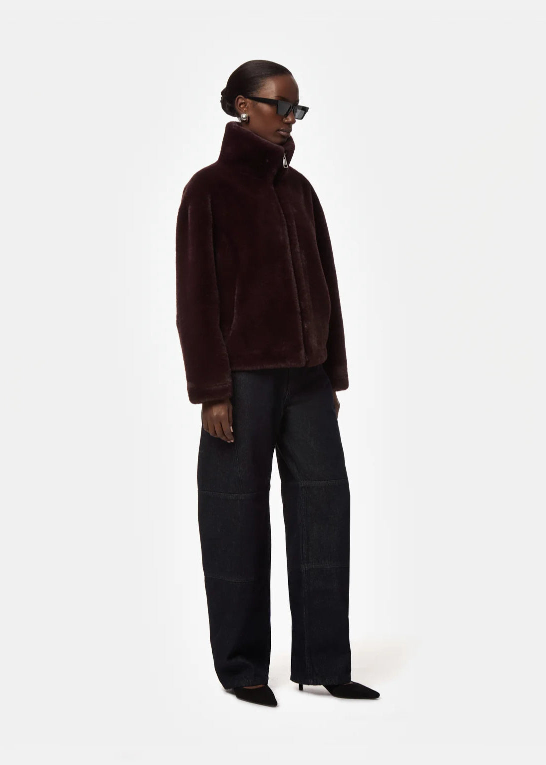A model wearing a dark brown plush faux-fur jacket with a high collar, paired with dark wide-leg trousers and black pointed shoes, photographed against a solid off-white background.