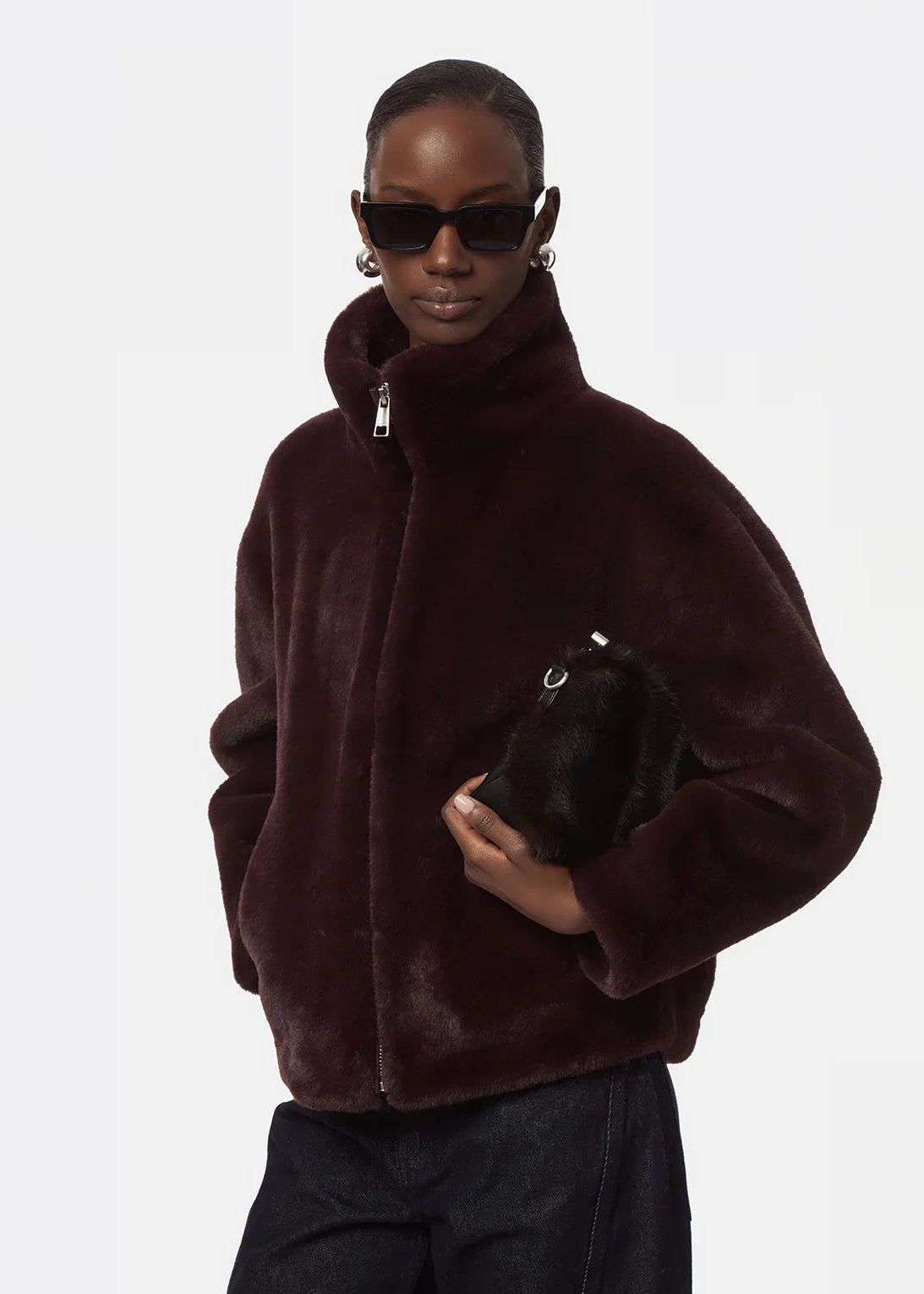 A model wearing a dark burgundy faux fur jacket, black sunglasses, and holding a matching dark fur clutch against a light gray background.