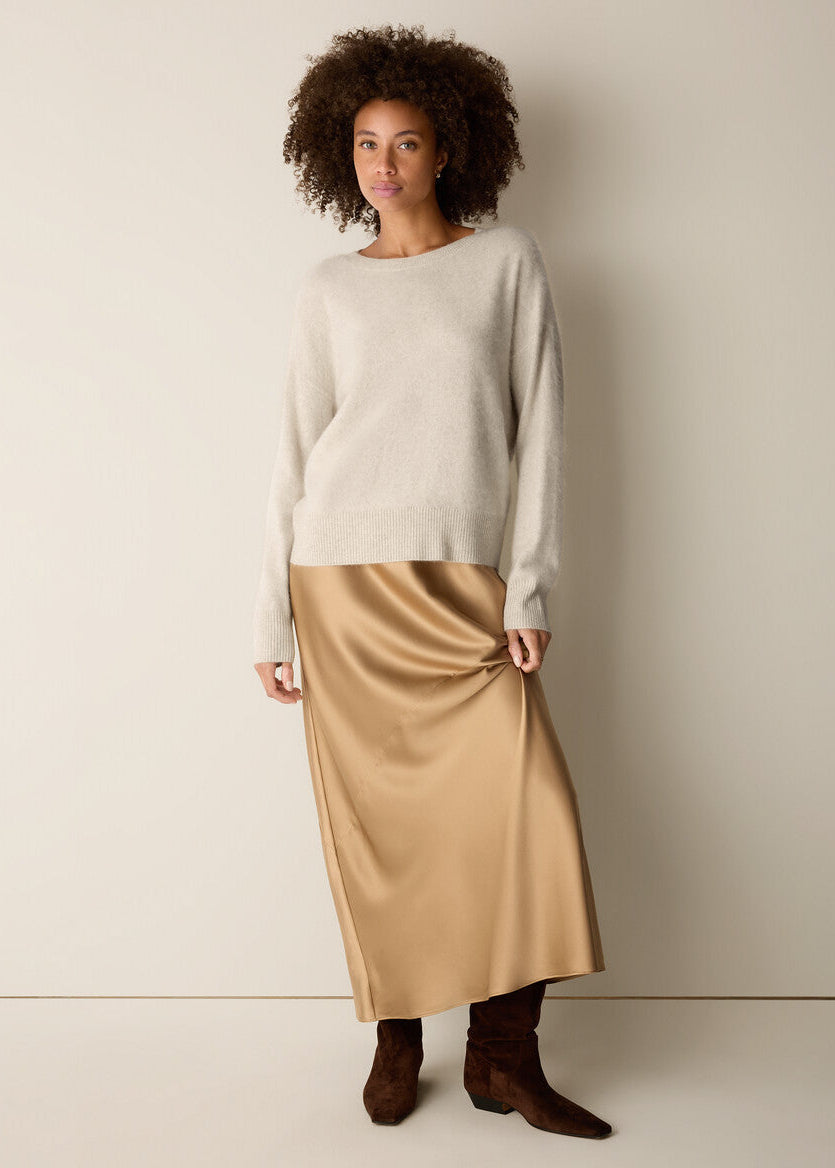 A woman stands against a plain light beige studio wall wearing a cream knit sweater, a satin-like tan midi skirt, and dark brown ankle boots.
