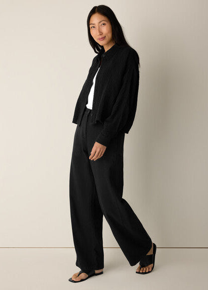 A woman modeling a black long-sleeve button-up shirt, black wide-leg trousers, a white top, and black slide sandals against a plain light beige studio background.
