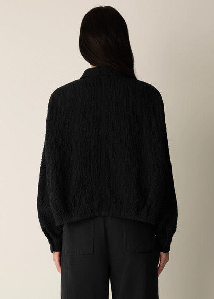 A model shown from the back wearing a black textured, crinkled long-sleeve jacket and black trousers against a plain, light beige background.