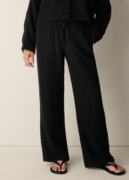 A person wearing black wide-leg pleated pants with a drawstring waist, paired with a matching black long-sleeve top and black flip-flop sandals, photographed against a plain light beige studio background.
