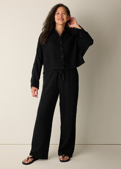 A woman modeling a black long-sleeve button-front top and matching wide-leg pants set, standing against a plain warm beige studio background.