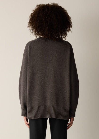 A person shown from the back wearing an oversized dark brown knit sweater and black pants against a plain light beige studio background.