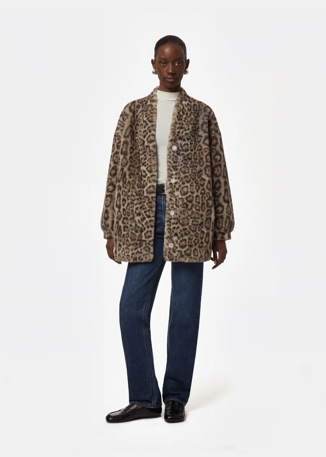 A model stands against a plain white studio background wearing a brown and beige leopard-print cardigan over a white turtleneck, blue jeans, and black leather shoes.