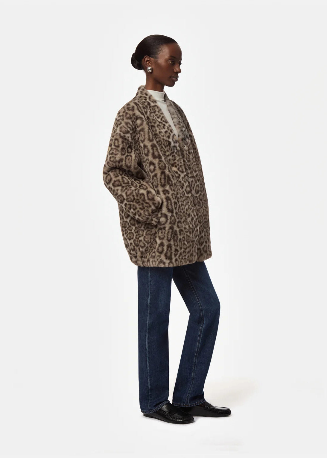 A model shown in profile wearing a brown and beige leopard-print faux fur jacket, dark blue jeans, and black shoes against a light gray studio background.