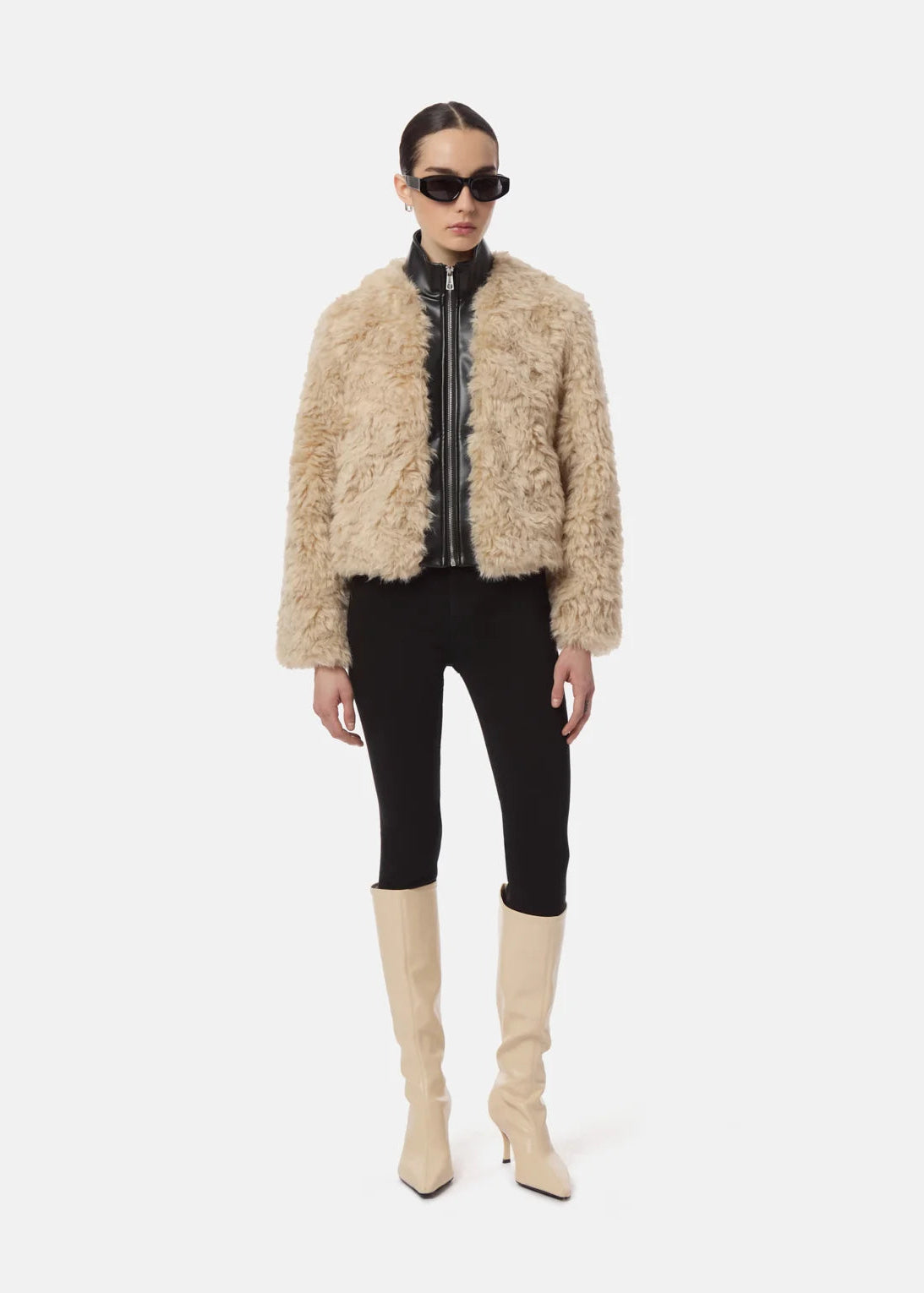 A model stands against a light gray studio background wearing a beige shaggy faux-fur cropped jacket over a black zip-front top, black pants, and beige knee-high heeled boots.