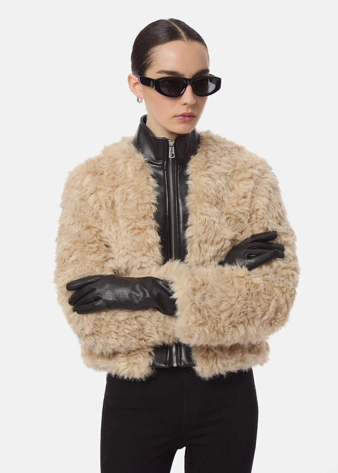 A model wearing a beige shaggy faux-fur cropped jacket with black leather trim, black leather gloves, and black pants, set against a plain light gray background.