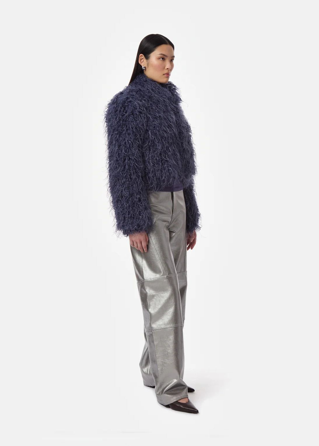 A model is shown from the side wearing a dark blue shaggy faux-fur cropped jacket, shiny silver wide-leg trousers, and black pointed flats, set against a plain light gray studio background.