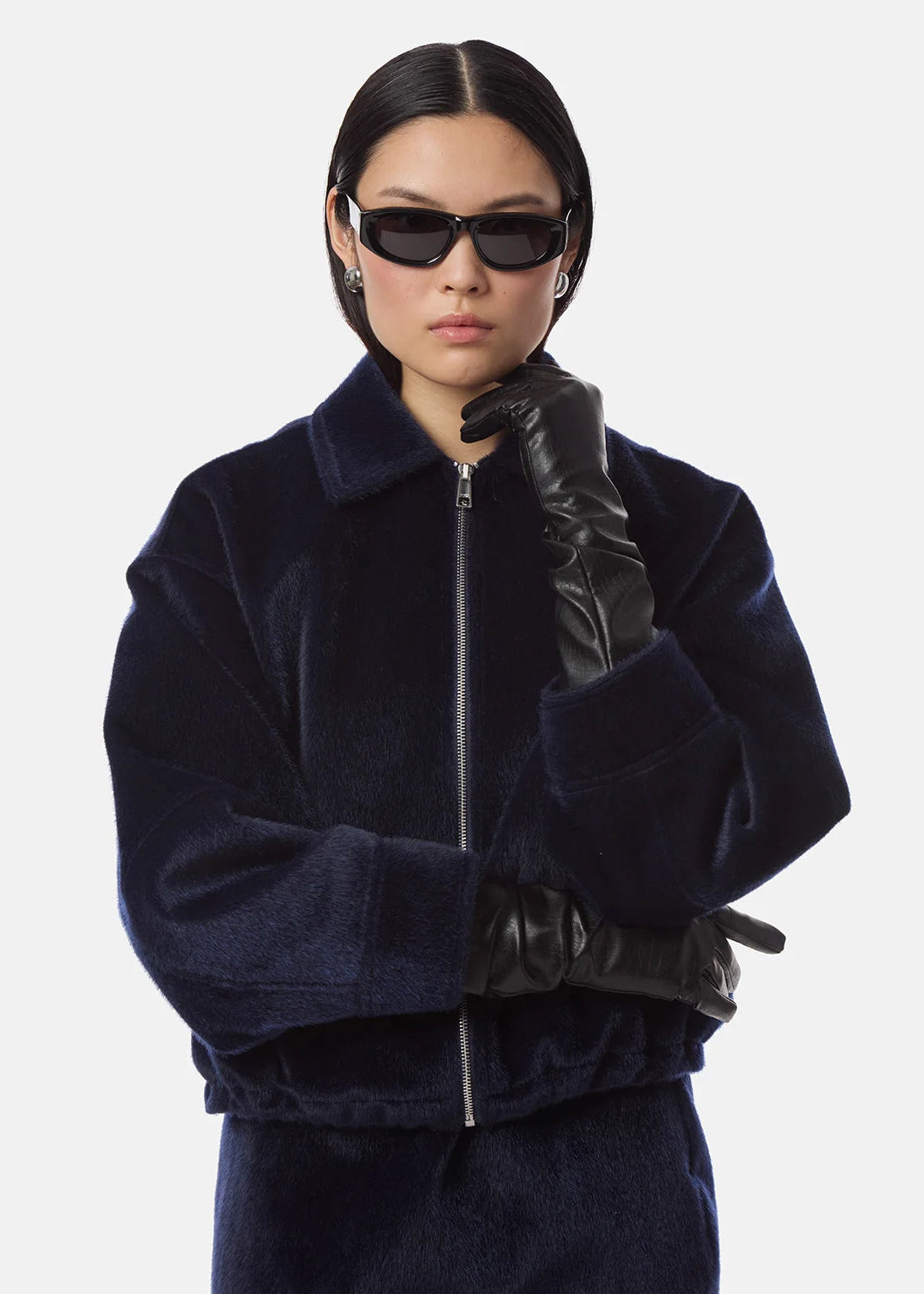 A model wearing a dark navy blue velvet jacket with a high collar, black leather gloves, and black sunglasses, posed against a light gray background.