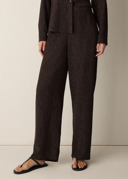 A model wearing dark brown textured wide-leg trousers and a matching dark brown long-sleeve top, styled with black thong sandals against a neutral beige studio background.