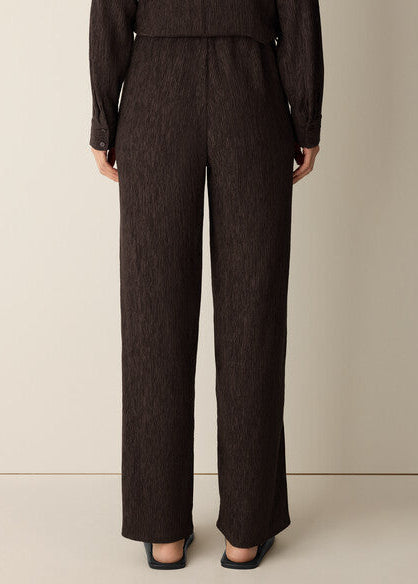 A model shown from the back wearing dark brown textured wide-leg trousers and a matching dark top, standing against a plain beige studio background.