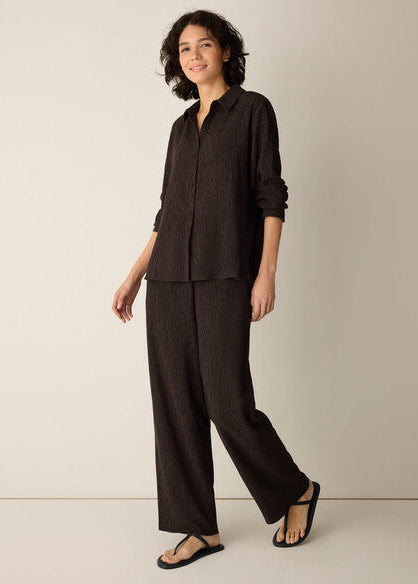 A woman modeling a dark brown linen two-piece set consisting of a long-sleeve button-up shirt and matching wide-leg trousers, paired with black sandals, against a plain beige studio background.