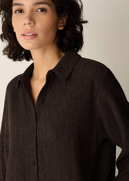 A close-up of a woman wearing a dark brown crinkled button-up shirt against a plain light beige background.