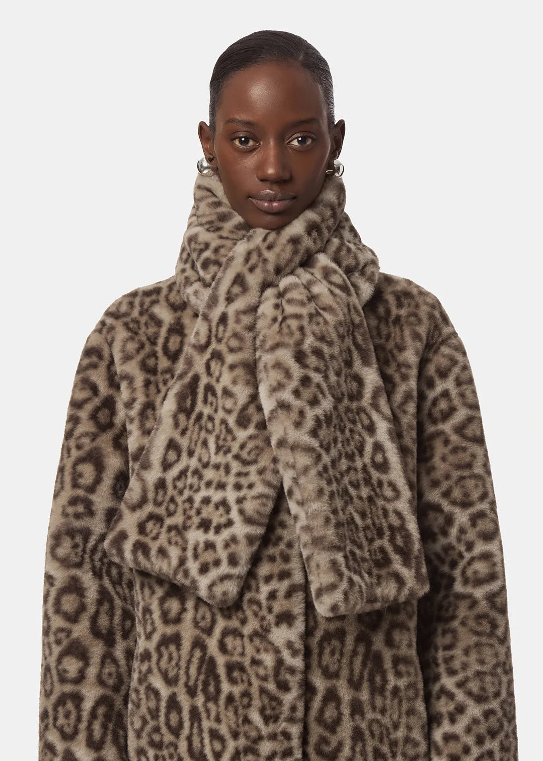 A model wearing a plush faux-fur coat and matching high-neck scarf in a brown and beige leopard print, photographed against a solid light gray background.
