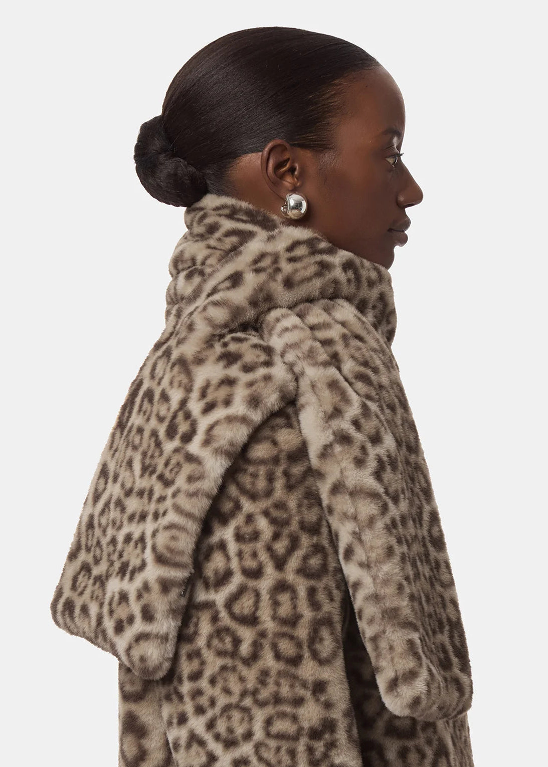 A side profile view of a model wearing a plush faux fur coat with a brown and beige leopard print pattern, set against a solid light gray background.
