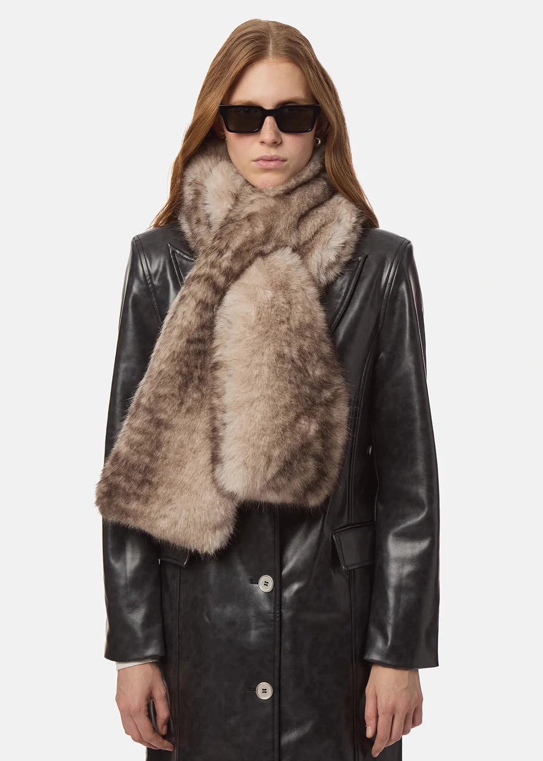 A model wearing a thick, shaggy faux-fur scarf in a mix of taupe and brown tones, styled over a black leather coat and black sunglasses, against a plain light gray background.