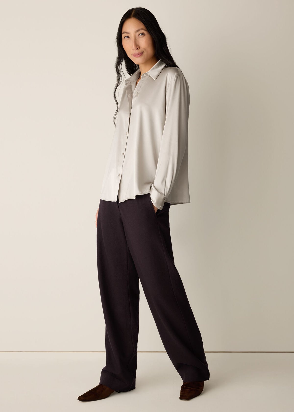 A woman modeling a light beige long-sleeve button-up shirt and dark charcoal wide-leg trousers, standing against a plain beige studio background.
