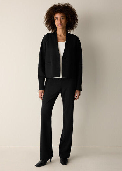 A model with curly dark hair stands against a plain beige studio wall wearing a black cropped cardigan, black wide-leg trousers, a white top, and black pointed shoes.