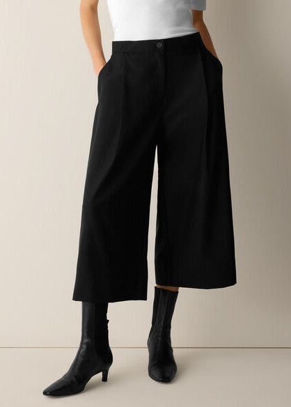 A model wearing black wide-leg cropped trousers with a button closure, paired with black pointed-toe ankle boots and a white top, standing against a neutral beige background.