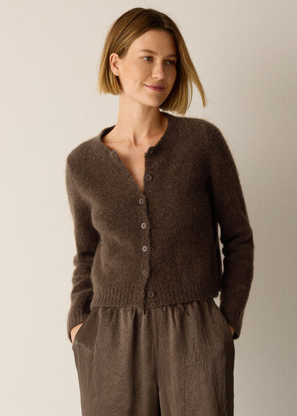 A woman wearing a brown fuzzy button-front cardigan with a split neckline and matching brown pants, standing against a plain light beige studio background.