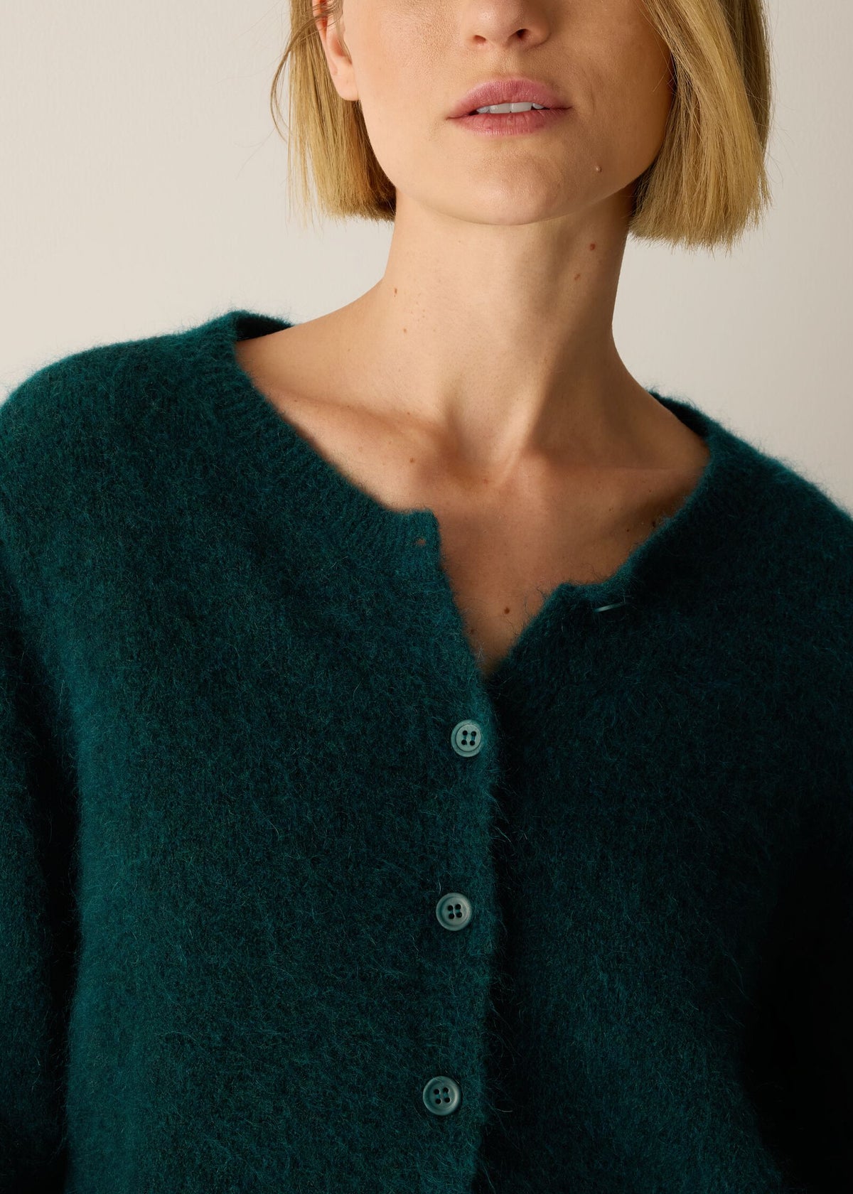 A close-up of a model wearing a dark teal fuzzy knit cardigan with a buttoned neckline.