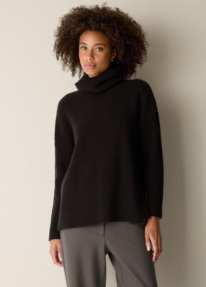 A woman with curly dark hair wearing a black ribbed turtleneck sweater and gray trousers, standing against a plain light beige wall.