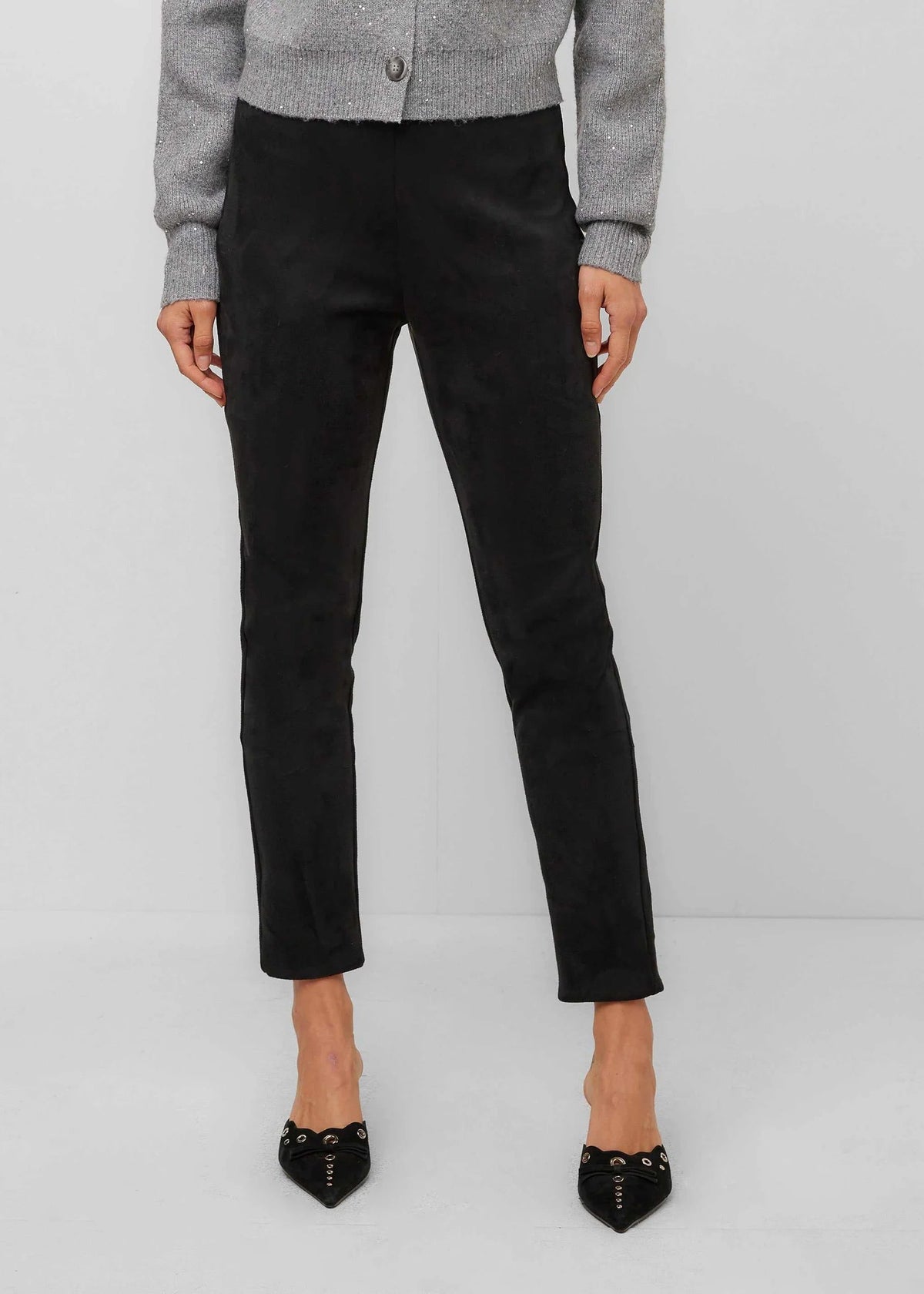 A model is shown from the waist down wearing black cropped ankle-length trousers, a gray cardigan, and black pointed-toe studded heels against a light gray studio background.
