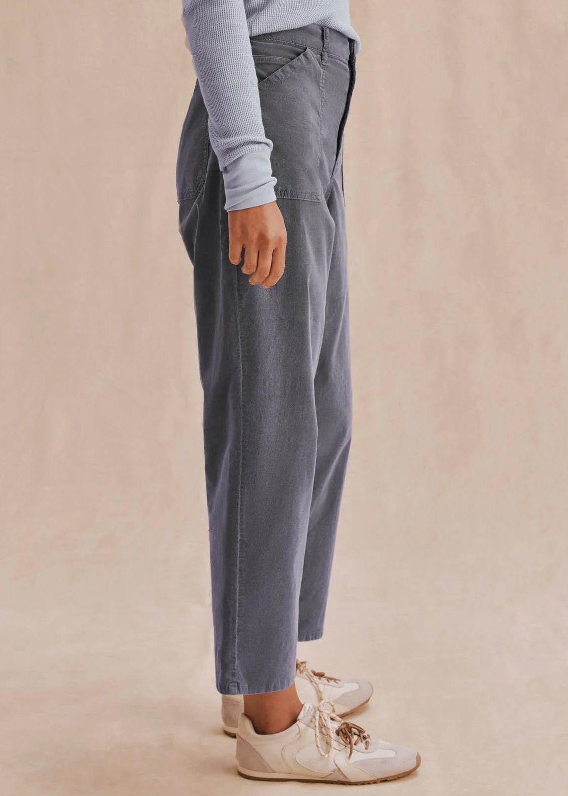 A person shown from the waist down wearing loose, wide-leg gray pants, a light blue long-sleeve top, and white sneakers against a soft beige studio backdrop.