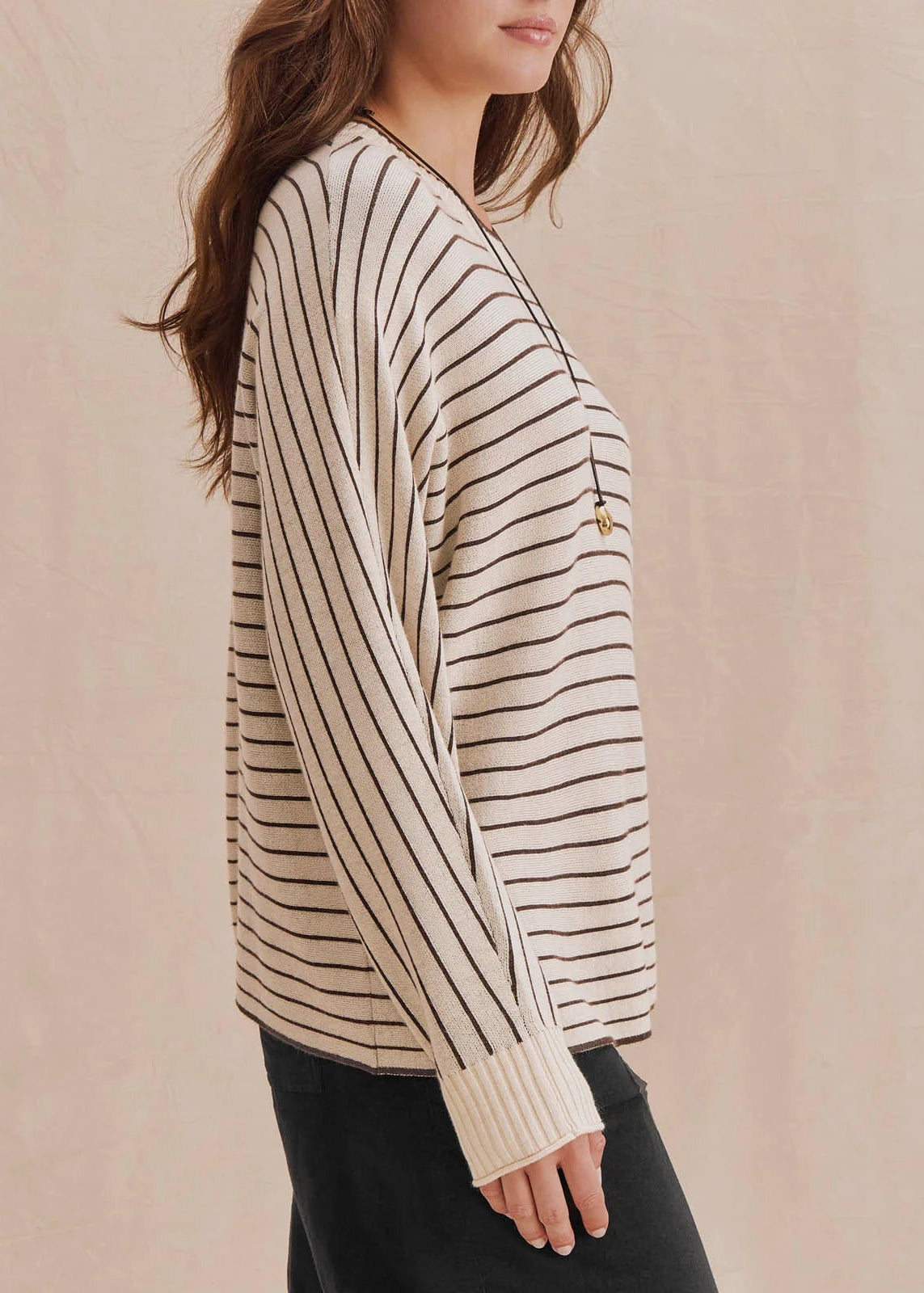 A woman wearing a cream-colored knit sweater with thin black vertical stripes and a small gold button detail on the chest, paired with black pants, set against a soft beige background.