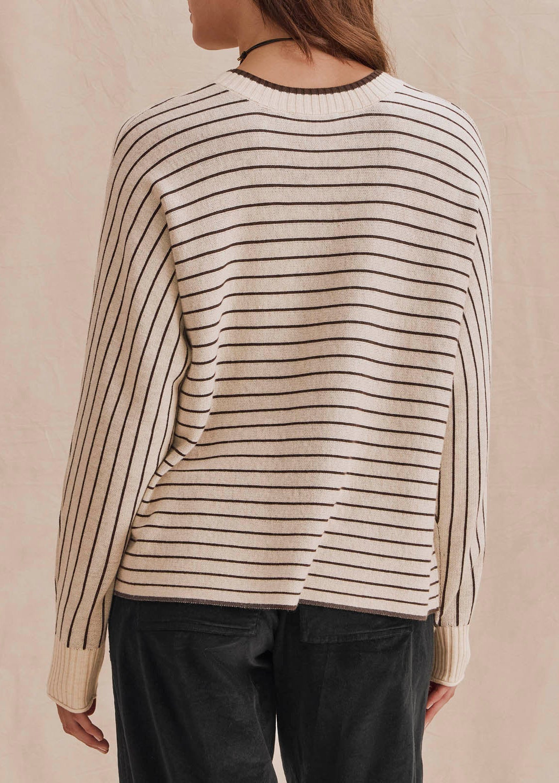 A person shown from the back wearing a cream knit sweater with thin dark brown horizontal stripes and dark pants, standing against a soft beige backdrop.