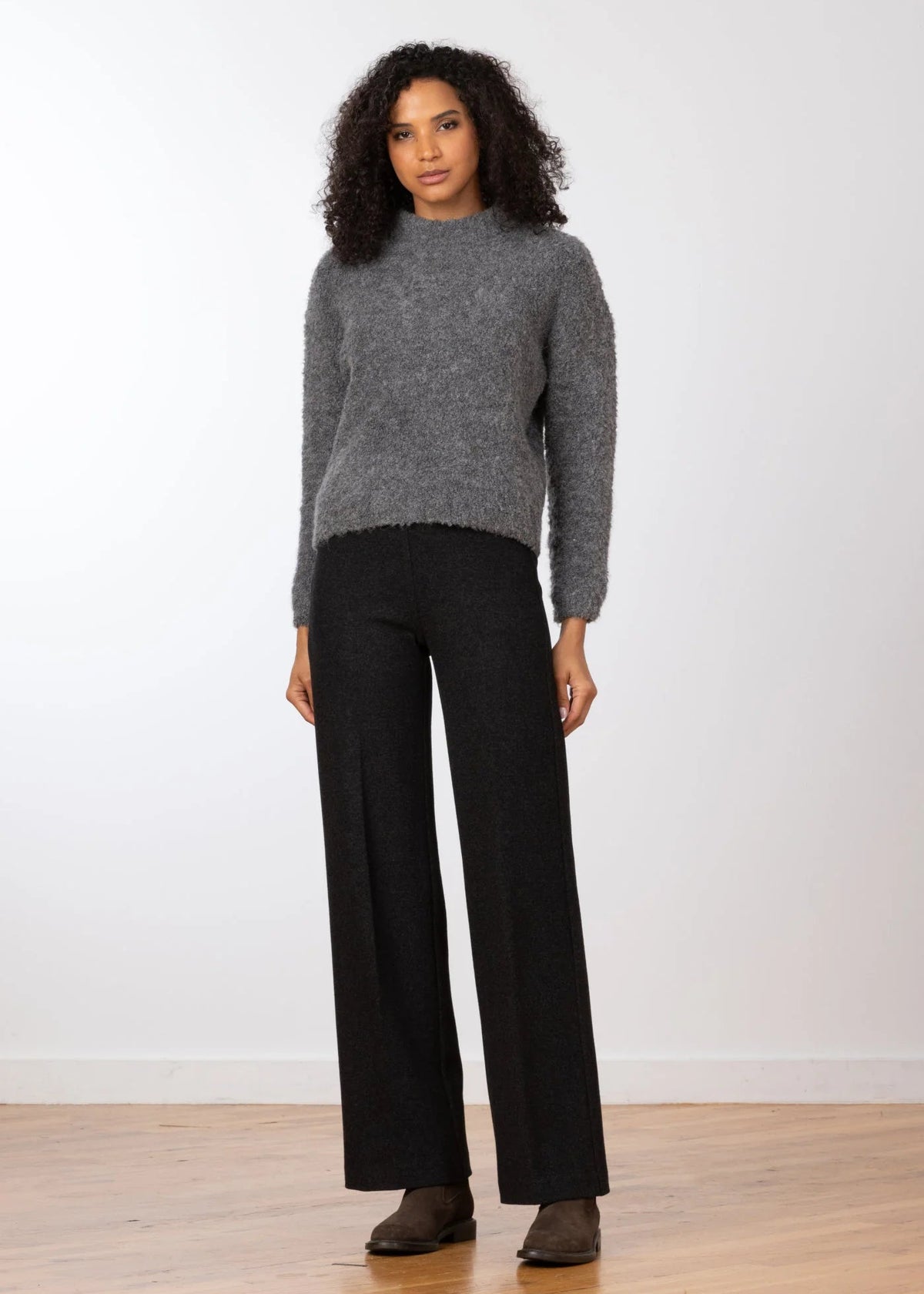 A woman modeling a gray fuzzy mock-neck sweater and black wide-leg trousers against a plain white wall and light wood floor.