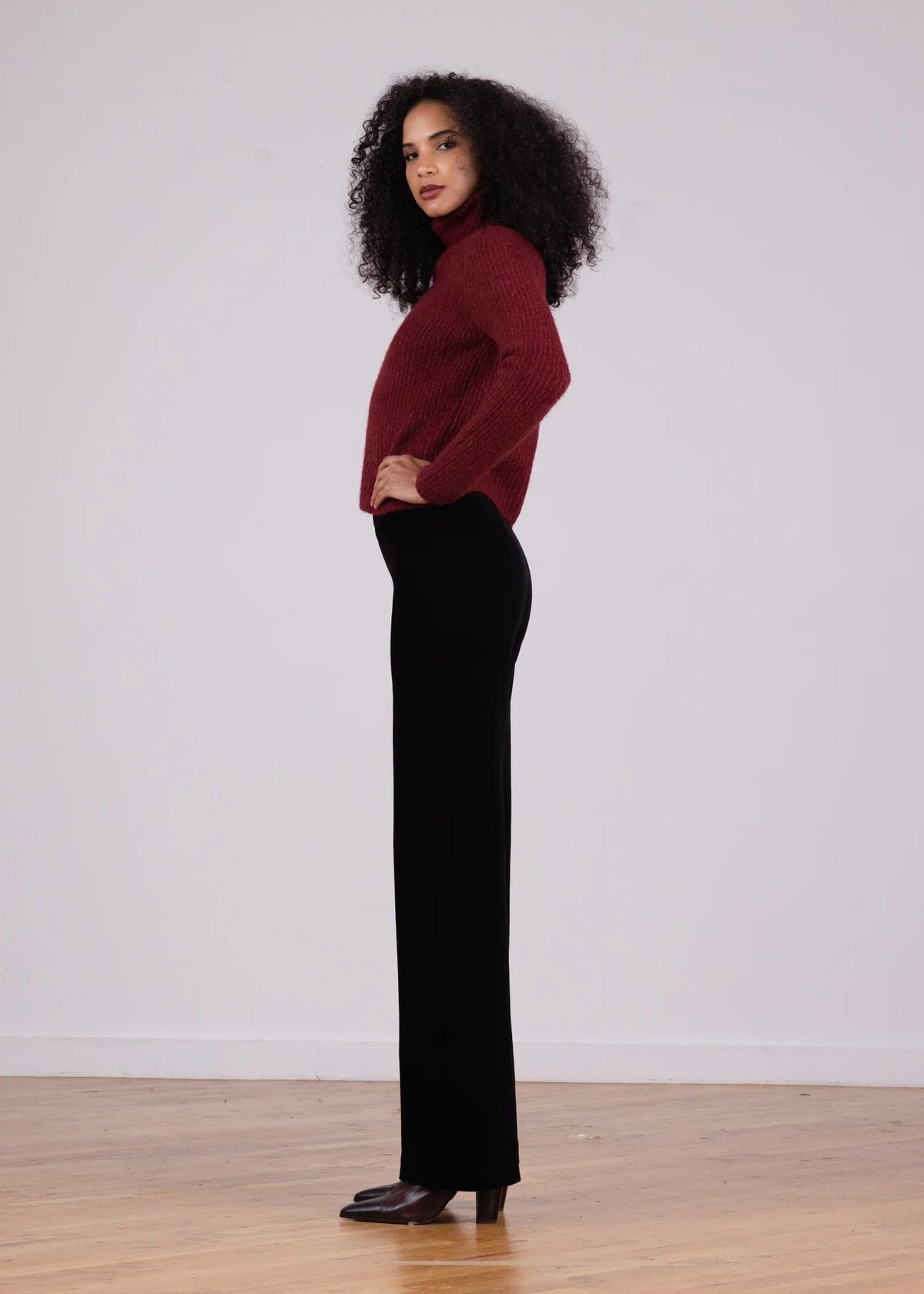 A woman stands in profile wearing a burgundy ribbed knit mock-neck sweater and black wide-leg pants, standing on a light wood floor against a plain white wall.