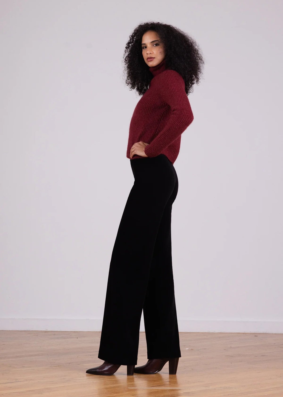 A woman stands in profile wearing a burgundy knit turtleneck sweater and black wide-leg pants, paired with dark brown heeled shoes, against a plain light gray wall and wooden floor.