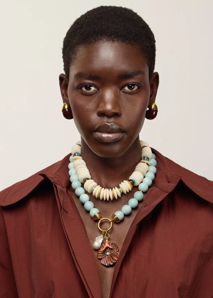 A close-up portrait of a model wearing a rust-brown collared shirt and layered necklaces, including a prominent beaded necklace with cream beads, turquoise-blue beads, and a gold-toned pendant, against a plain light beige background.