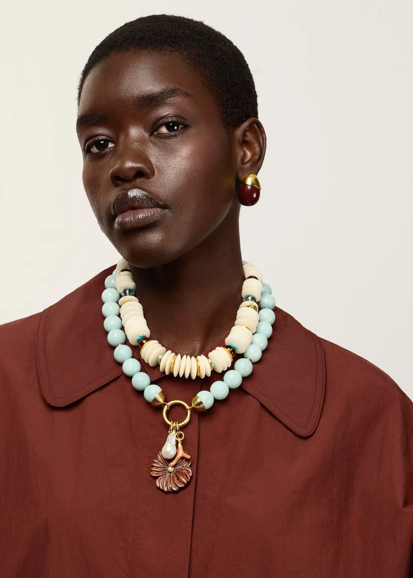 A close-up of a model wearing a multi-strand statement necklace with light blue beads, cream bone-like beads, and a gold shell pendant, paired with a rust-brown collared shirt.