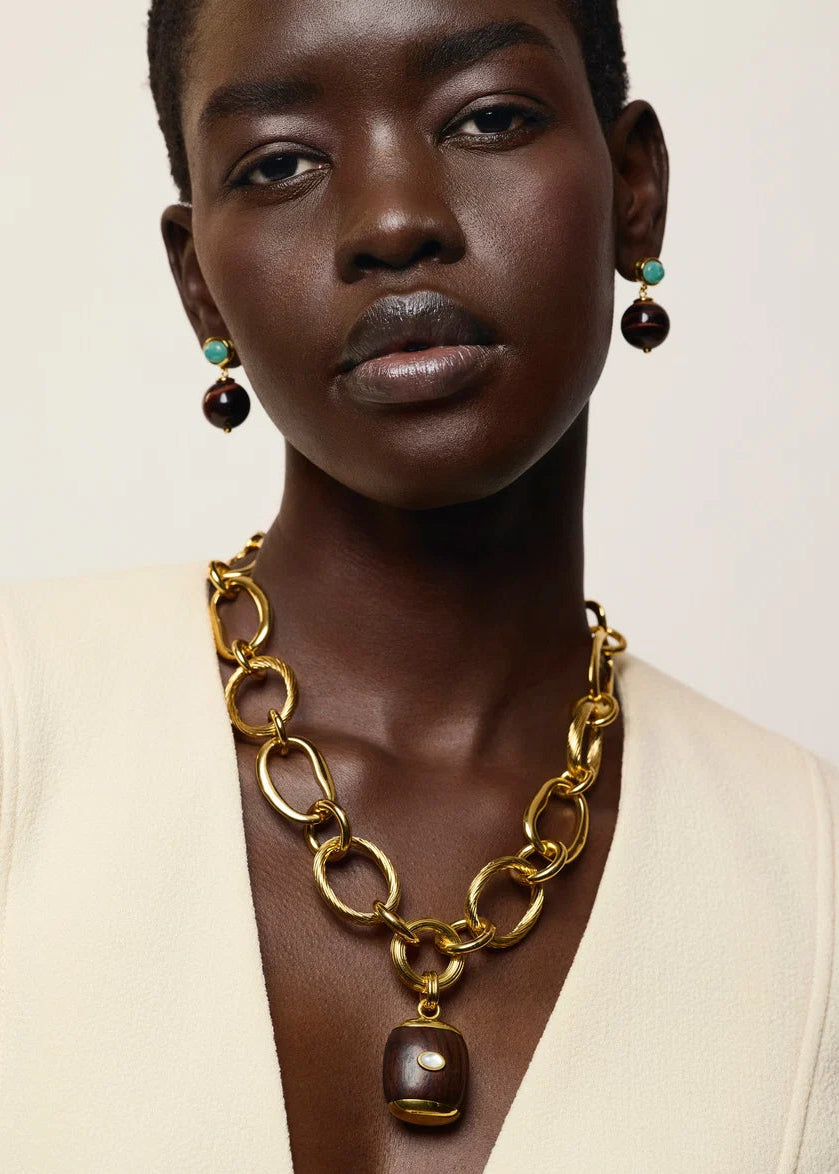 A close-up of a model wearing a chunky gold chain necklace with a dark brown pendant and matching drop earrings with turquoise and dark brown beads, paired with a cream-colored top.