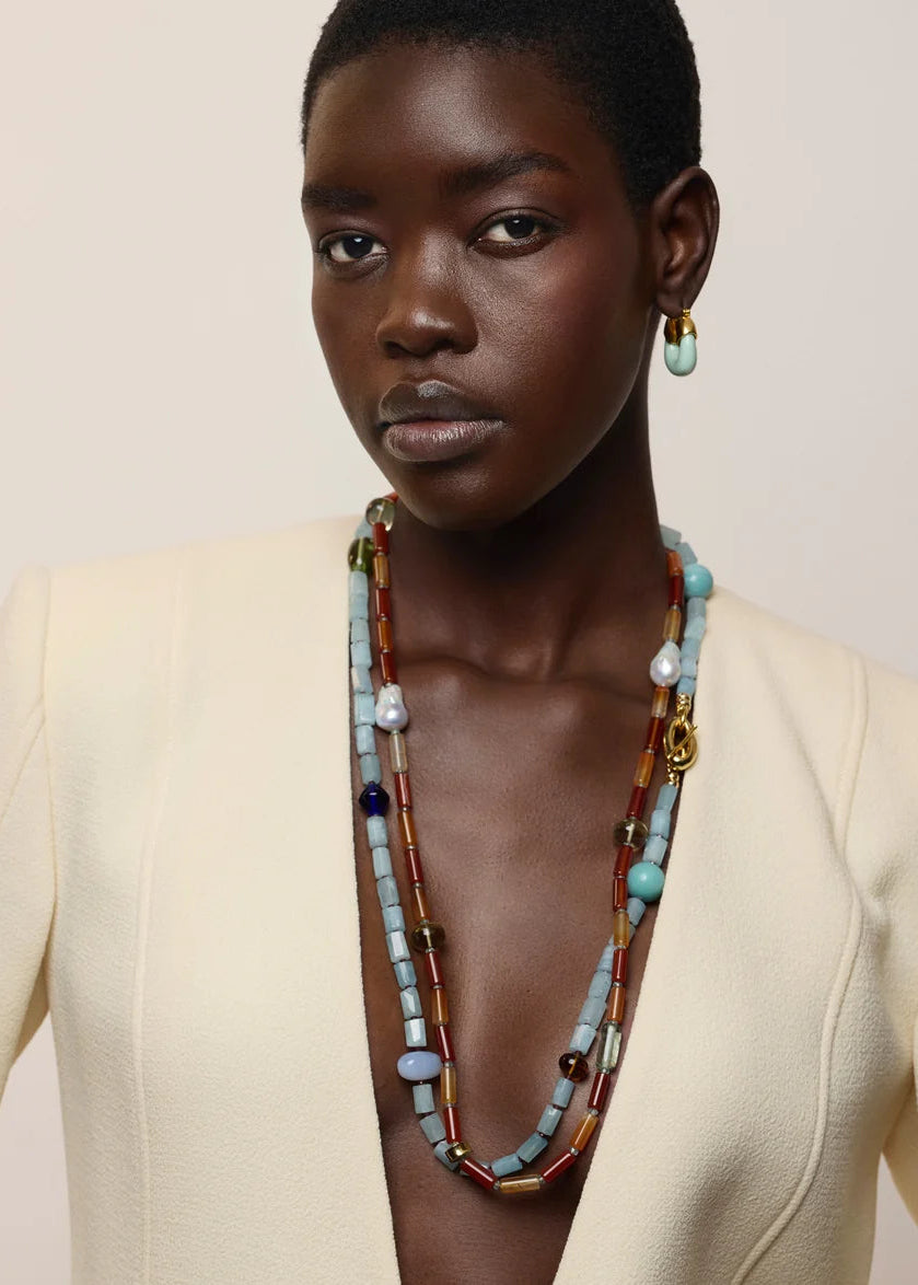 A model wearing a cream blazer and a long, multi-strand beaded necklace with light blue, white, and brown beads, paired with a matching light blue teardrop earring.