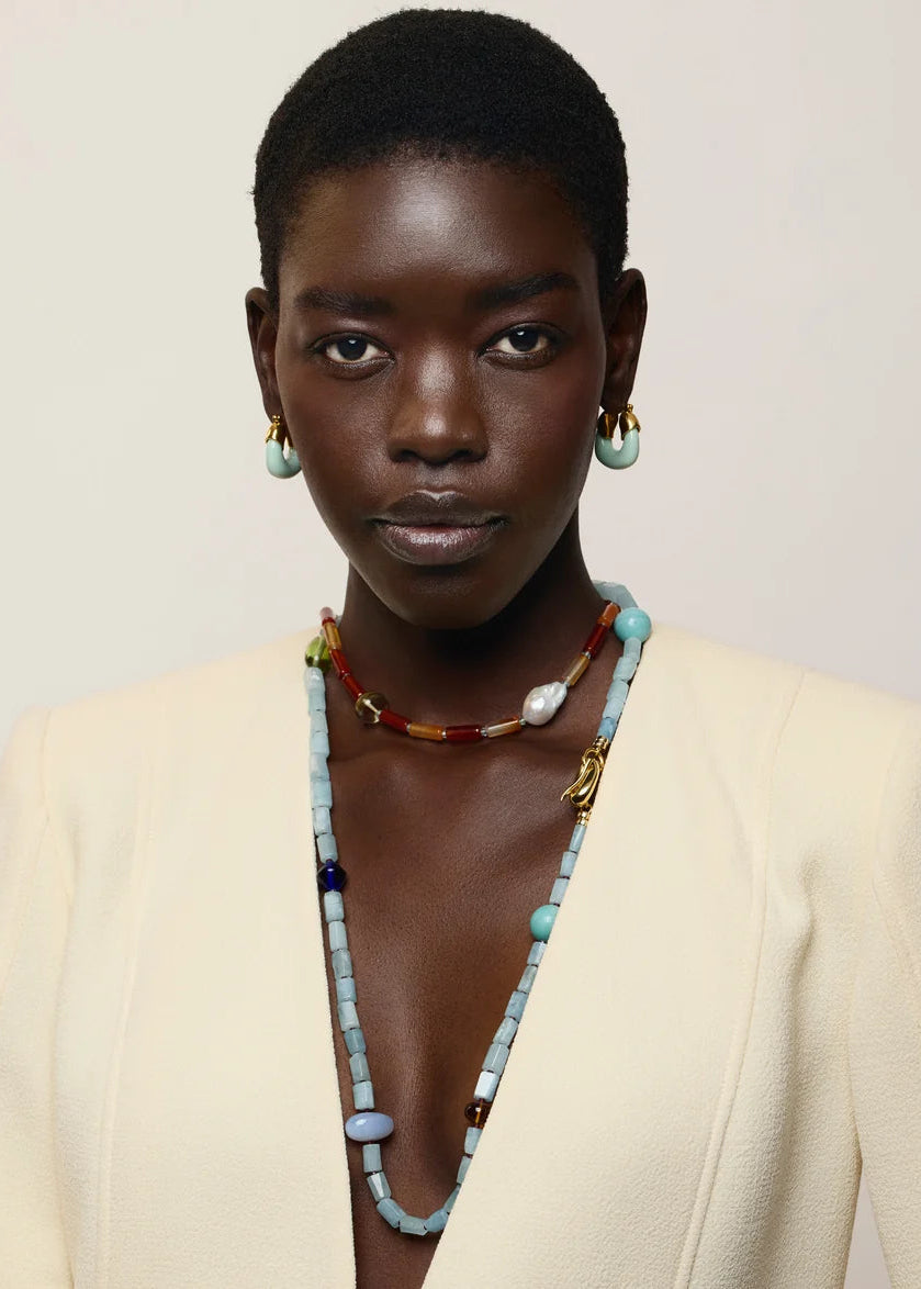 A model with short dark hair is shown from the chest up, wearing a cream-colored blazer and layered beaded necklaces and earrings. The jewelry features light blue, white, and brown beads with gold-toned metal accents.