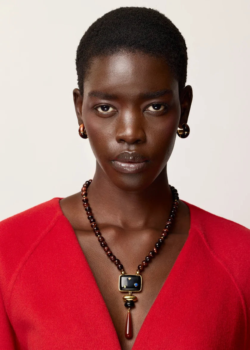 A model with short dark hair wearing a vibrant red sweater, a dark beaded necklace with a rectangular black and gold pendant, and matching gold hoop earrings, set against a plain off-white background.