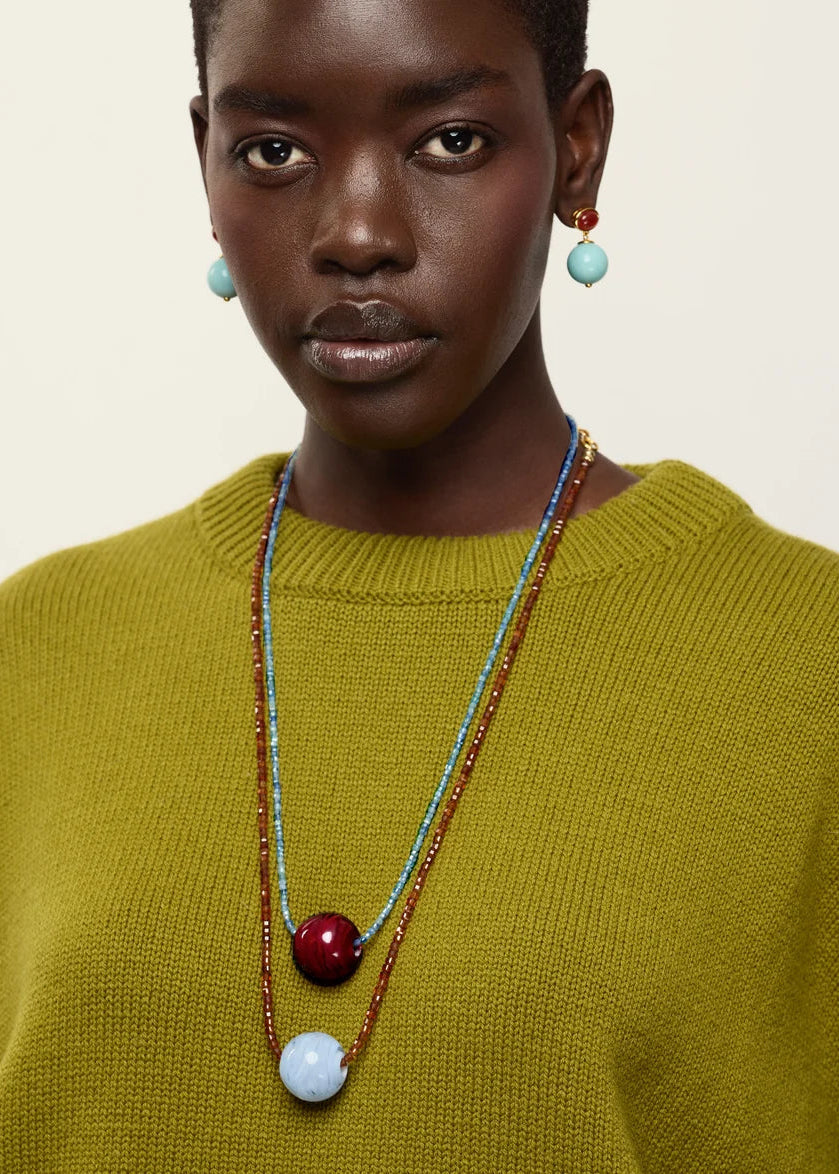 A close-up of a model wearing a multi-strand beaded necklace with light blue, dark red, and brown beads, along with matching light blue drop earrings, paired with a textured olive green sweater against a plain off-white background.