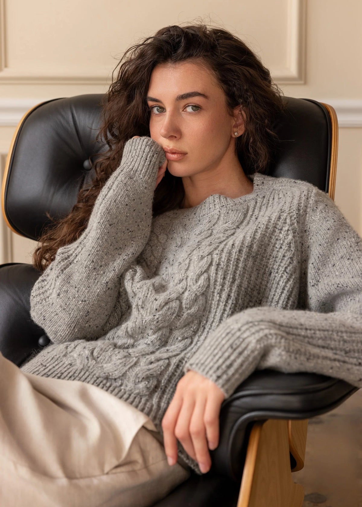A woman with long, wavy brown hair sits in a black leather and wood lounge chair, wearing a chunky light gray cable-knit sweater and beige trousers.