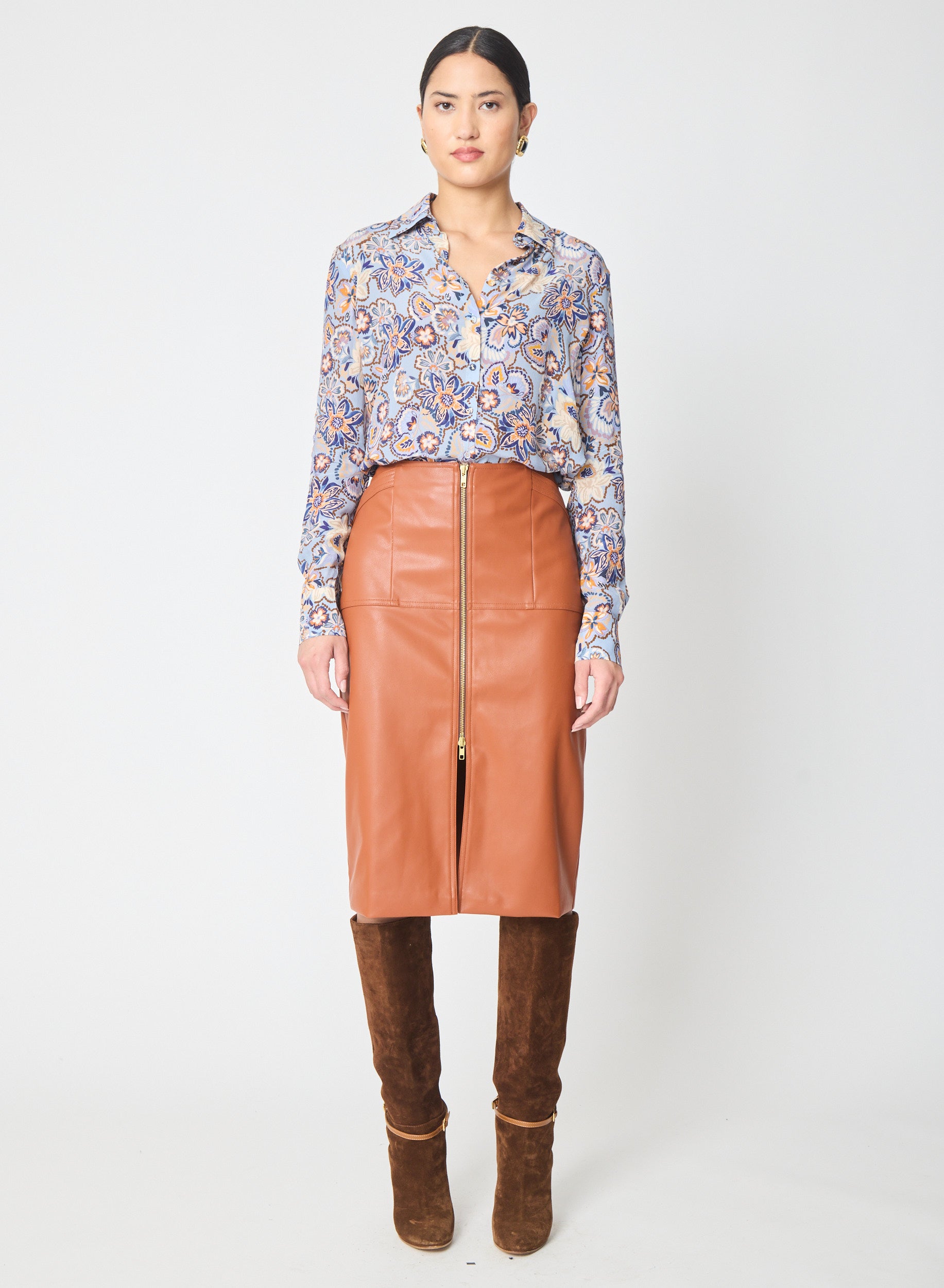 A model stands against a light gray studio background wearing a long-sleeve button-up shirt with a blue, purple, and gold paisley print, a tan faux-leather pencil skirt with a front zipper, and brown suede knee-high boots.