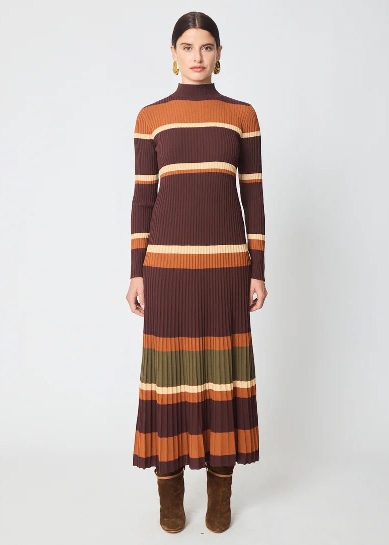 A woman stands against a light gray studio background wearing a long-sleeve ribbed knit mock-neck midi dress with horizontal stripes in dark brown, rust orange, olive green, and cream, paired with brown suede ankle boots.