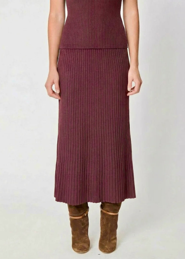 A model wearing a ribbed knit midi skirt in a deep plum purple color, paired with brown suede ankle boots, against a plain light gray background.