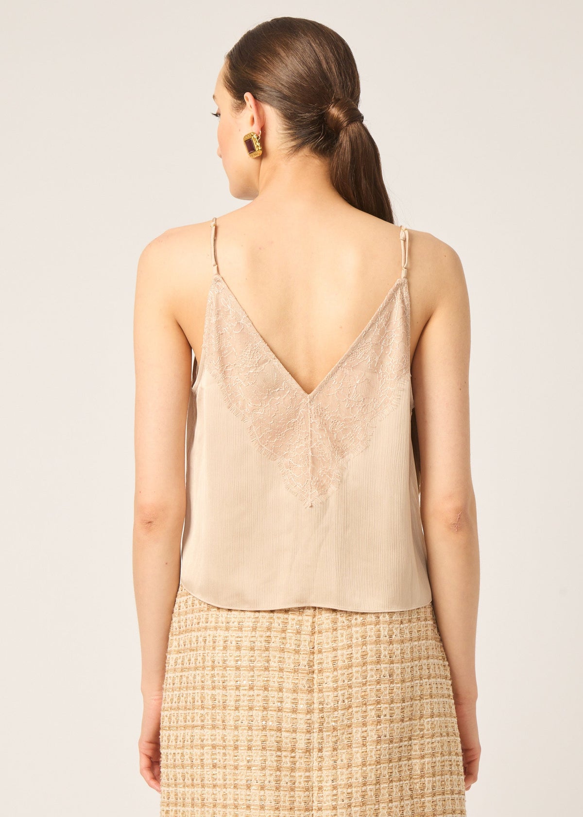A woman shown from the back wearing a light beige spaghetti-strap camisole top with a deep V-neckline and a textured woven skirt, against a plain light beige studio background.