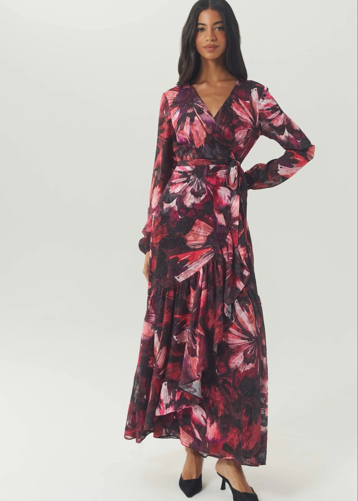 A woman modeling a long-sleeved, wrap-style maxi dress with a dark floral print in shades of red, pink, and black, paired with black pointed-toe heels, against a plain light gray studio background.